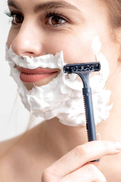 Красива молода біла жінка голить обличчя бритвою на білому тлі. Красива жінка з піною для гоління на обличчі
 - Фото, зображення
