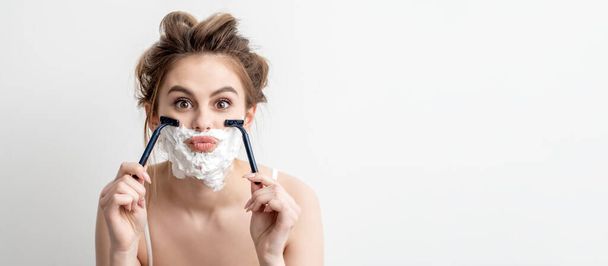 Beautiful young caucasian woman shaving her face by two razors on white background. Pretty woman with shaving foam on her face - Photo, Image