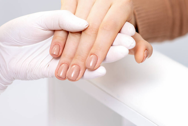 Close-up van de menselijke hand in beschermende handschoen houden vrouwelijke vingers met beige manicure in schoonheidssalon - Foto, afbeelding