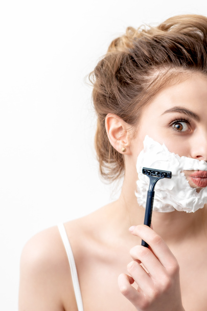 Beautiful young caucasian woman shaving her face by razor on white background. Pretty woman with shaving foam on her face - Φωτογραφία, εικόνα