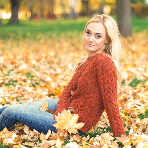 Belle jeune femme caucasienne en pull rouge assise sur les feuilles dans le parc d'automne - Photo, image