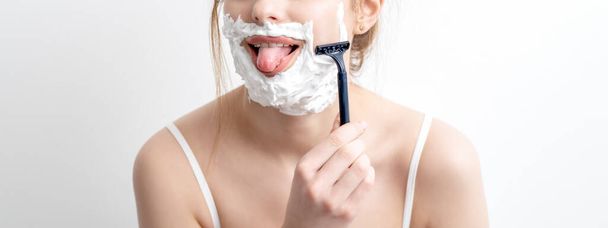 Beautiful young caucasian woman shaving her face by razor on white background. Pretty woman with shaving foam on her face - Фото, изображение