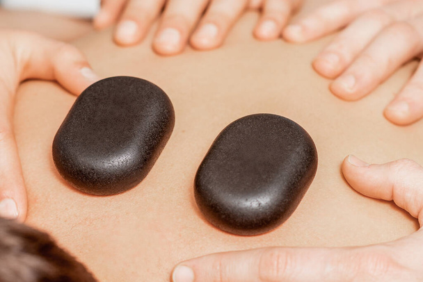 Τα χέρια των θεραπευτών μασάζ κάνει μασάζ στην πλάτη, ενώ ζεστό πέτρες στο πίσω μέρος του ανθρώπου κοντά σε spa. - Φωτογραφία, εικόνα