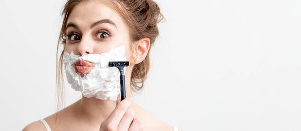 Красива молода біла жінка голить обличчя бритвою на білому тлі. Красива жінка з піною для гоління на обличчі
 - Фото, зображення