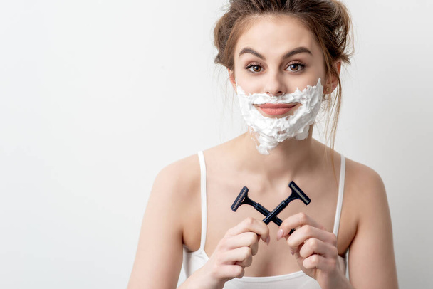 Beautiful young caucasian woman with shaving foam on her face and two razors in her hands on white background - Photo, Image
