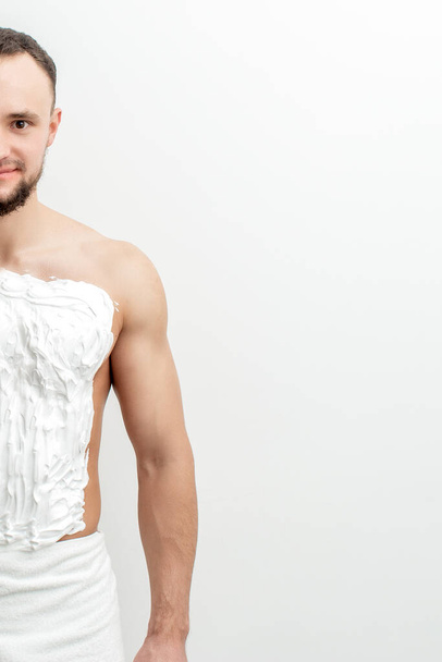 Young caucasian man with beard holds razor shaves his chest with white shaving foam on white background. Man shaving his torso - 写真・画像
