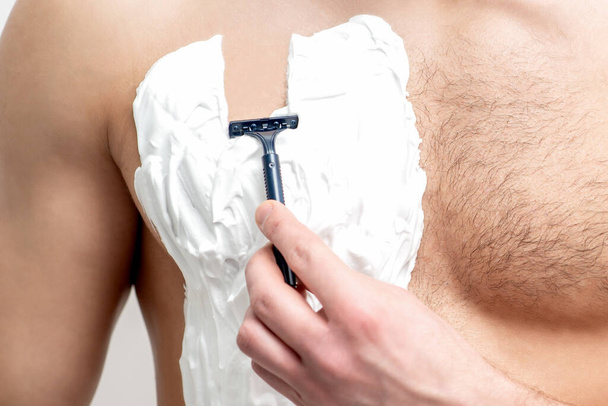 Jonge blanke man met baard houdt scheermes scheert zijn borst met wit scheerschuim op witte achtergrond. Man scheert zijn romp - Foto, afbeelding