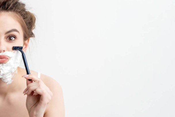 Schöne junge kaukasische Frau rasiert ihr Gesicht durch zwei Rasiermesser auf weißem Hintergrund. Hübsche Frau mit Rasierschaum im Gesicht - Foto, Bild