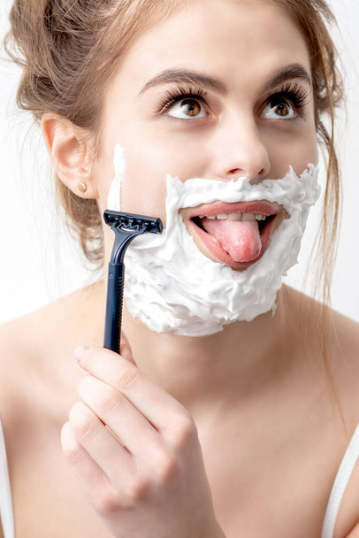 Hermosa mujer caucásica joven afeitándose la cara con una navaja de afeitar sobre fondo blanco. Mujer bonita con espuma de afeitar en la cara - Foto, imagen