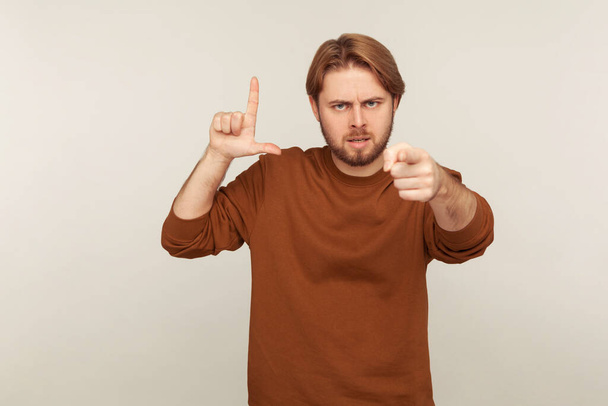 You're loser! Portrait of angry bossy man with beard in sweatshirt showing loser gesture and pointing finger to camera, blaming for mistake and dismissing from job. indoor studio shot isolated  - Фото, зображення