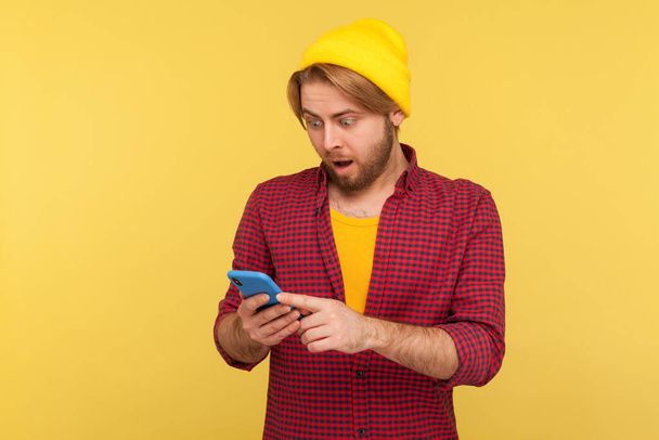 Amazed hipster guy in beanie hat and checkered shirt looking surprised at cell phone, reading shocking message, chatting in social network, astonished by mobile app. indoor studio shot isolated  - Foto, Imagem