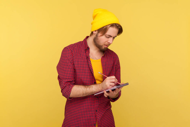 Hipster trendy guy in beanie hat looking inspired by creative idea, eager writing thought reminder to-do list into notebook, taking notes in paper, business plan. indoor studio shot isolated on yellow  - 写真・画像