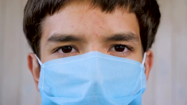 Lähellä kasvot nuori mies yllään sininen lääketieteellinen naamio ja katsot kameraa. Potilaan muotokuva suojapeitteessä, poika estää covid-19-koronaviruksen influenssapandemian sisätiloissa - Materiaali, video