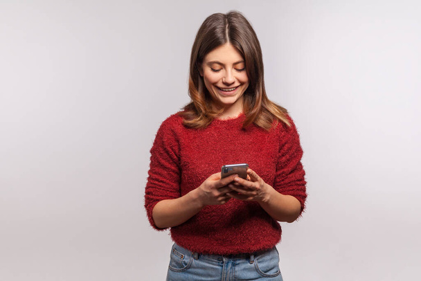 Happy brunette girl in shaggy sweater using mobile phone and smiling laughing joyfully, typing or reading funny message on social network, online communication. studio shot isolated on gray background  - Фото, изображение