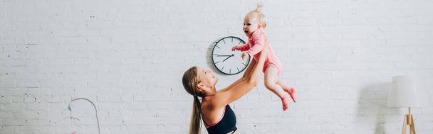 Panoramic shot of woman in sportswear holding infant daughter at home  - Фото, изображение