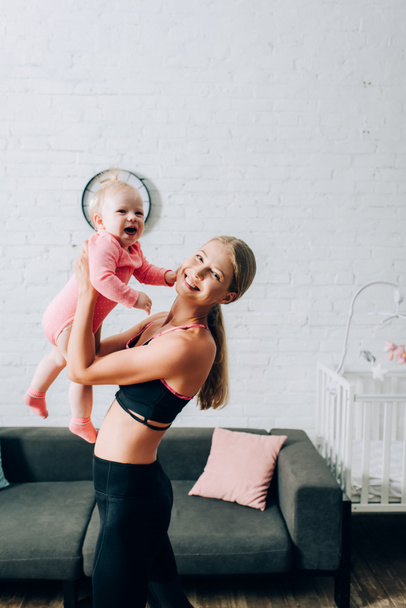 Woman in sportswear looking at camera while holding daughter in living room - Foto, imagen