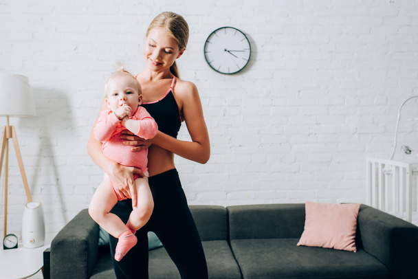 Fit woman in sportswear holding baby daughter on hands in living room  - 写真・画像