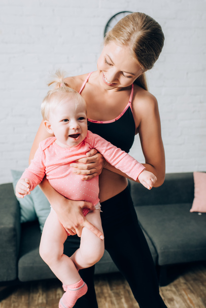 Mère en vêtements de sport tenant bébé fille dans le salon  - Photo, image