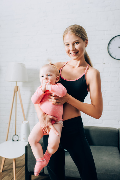 Fitt nő sportruházatban tartja csecsemő gyermek a nappaliban  - Fotó, kép