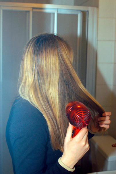 Genç bir kadın uzun sarı saçlarını kızıl saç kurutma makinesiyle kurutuyor. - Fotoğraf, Görsel