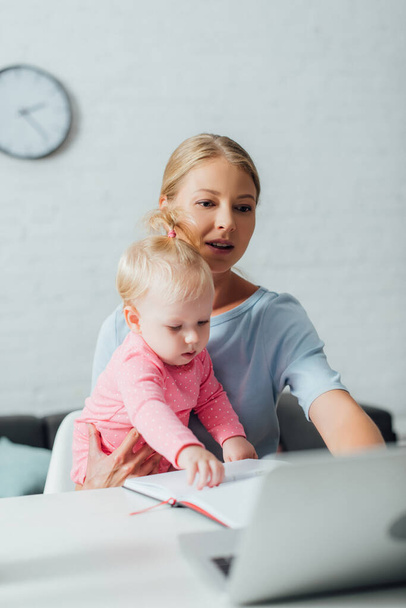 A laptopot használó nő szelektív fókusza, miközben a baba lányát tartja otthon - Fotó, kép