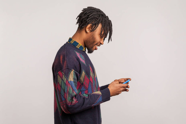 Profile portrait of happy trendy african guy with dreadlocks in sweatshirt using smartphone and smiling, pleased satisfied with connection quality. Indoor studio shot isolated on gray background  - Foto, imagen
