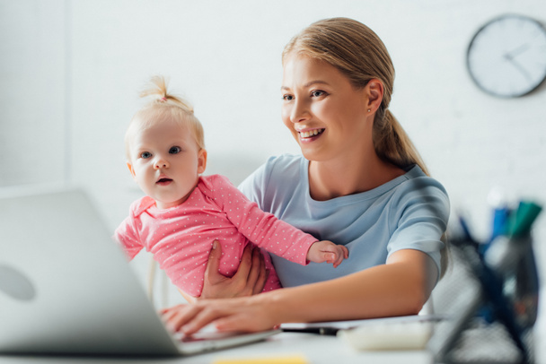 Selective focus of mother holding baby girl and using laptop at home  - Foto, Imagem
