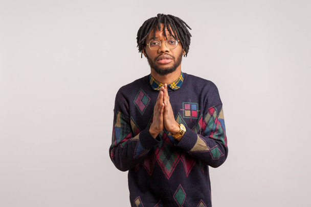 Concerned upset bearded african guy with dreadlocks in casual sweatshirt and eyeglasses holding hands in pray, asking for help and support, human rights. Indoor studio shot isolated on gray background  - Photo, image