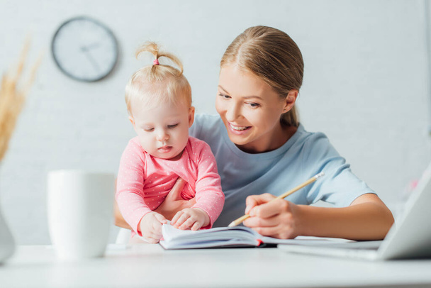 Concentration sélective de la femme écrivant sur le carnet et tenant bébé fille tout en travaillant à la table  - Photo, image