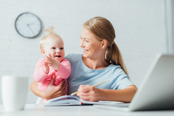 Focus selettivo della madre che guarda la bambina mentre lavora con notebook e laptop a casa  - Foto, immagini