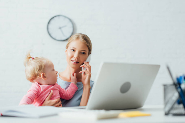 Selective focus of woman talking on smartphone while holding baby girl near laptop and stationery on table  - Foto, immagini