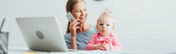 Panoramiczne ujęcie kobiety rozmawiającej przez telefon komórkowy i trzymającej córkę przy laptopie na stole  - Zdjęcie, obraz