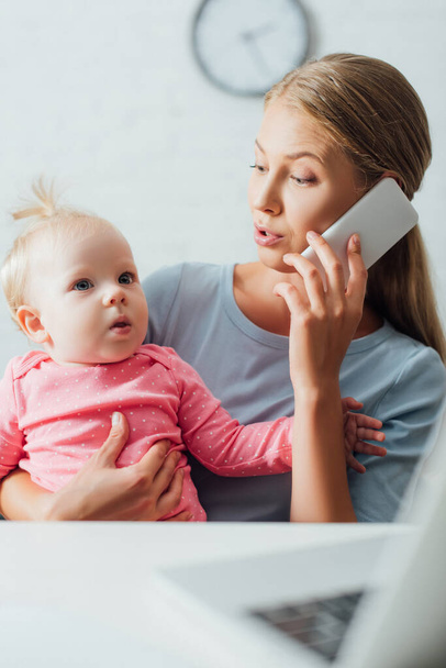 Focus selettivo della madre che tiene in braccio la bambina e parla sullo smartphone mentre lavora a casa  - Foto, immagini