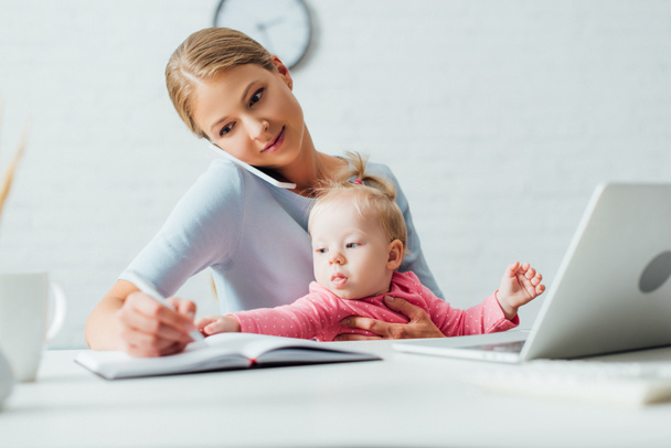 Selective focus of infant girl sitting near mother talking on smartphone and writing on notebook at home  - Фото, изображение