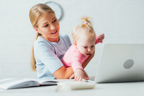 Selectieve focus van de vrouw die baby dochter terwijl het werken met notebook en laptop thuis  - Foto, afbeelding