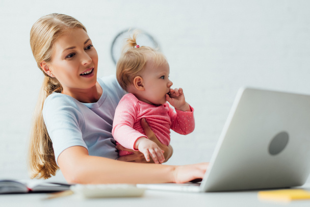 Selective focus of mother holding baby girl while working on laptop at home  - Foto, Imagem