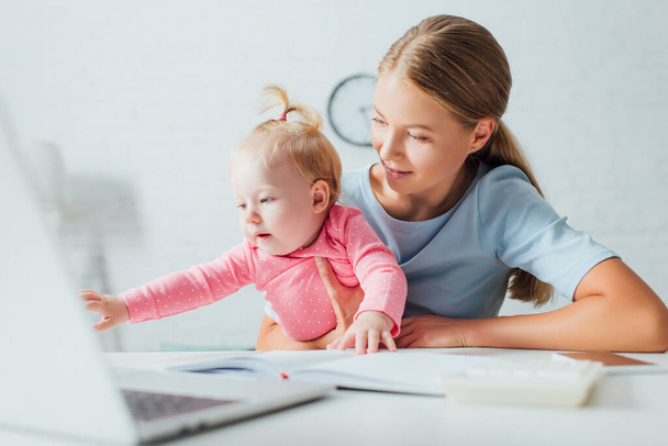 Selective focus of woman holding baby girl on hands near notebook and gadgets on table  - Фото, зображення