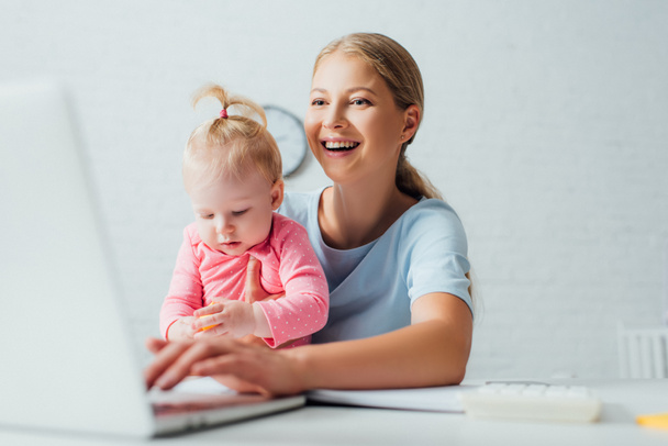 Selective focus of woman holding daughter and using laptop at table  - Foto, Imagen