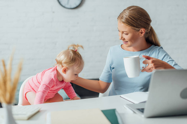 Selective focus of woman holding cup and touching baby girl near laptop and stationery on table  - Foto, Bild