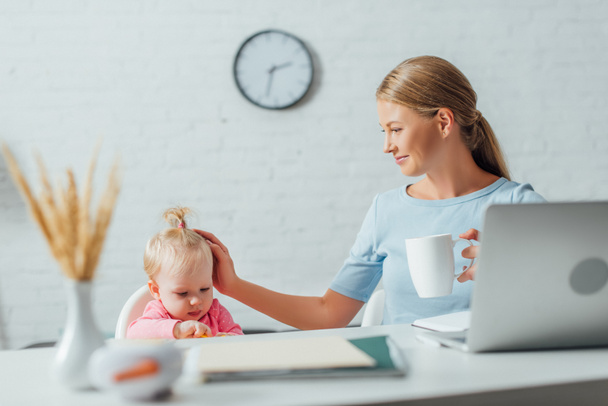 Selective focus of freelancer with cup touching infant daughter near laptop and stationery on table  - Photo, Image