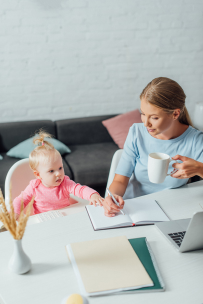 Selective focus of baby girl sitting near mother holding cup and writing on notebook at home  - Valokuva, kuva