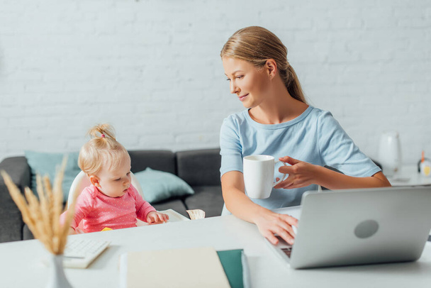 Enfoque selectivo de la mujer sosteniendo la taza y usando el ordenador portátil cerca de la niña en casa  - Foto, Imagen