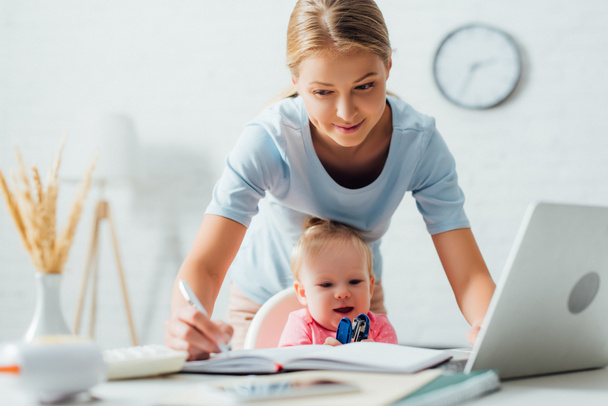Selektivní zaměření ženy psaní na notebook při práci v blízkosti kojence se sešívačkou u stolu  - Fotografie, Obrázek