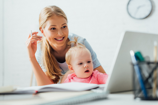 Selective focus of woman holding pen and looking at laptop near child at home  - Foto, Imagem