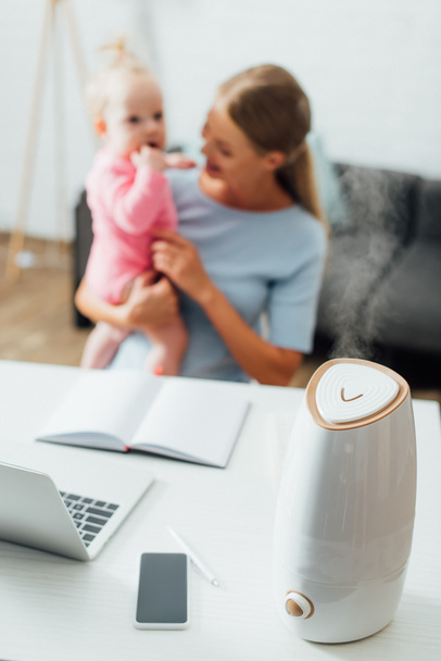 Selektiver Fokus des Luftbefeuchters in der Nähe von Gadgets auf dem Tisch und Mutter hält Säugling im Hintergrund  - Foto, Bild