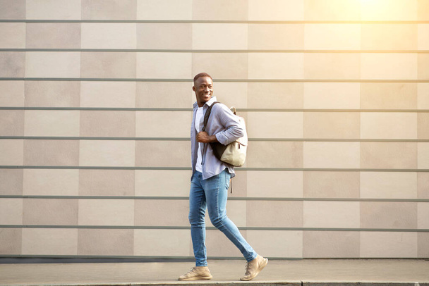 Junger Schwarzer in Freizeitkleidung mit Rucksack läuft nahe Ziegelmauer auf der Stadtstraße - Foto, Bild