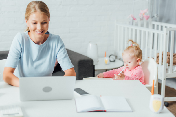 Selective focus of baby girl holding pen while mother using laptop at home  - Фото, изображение