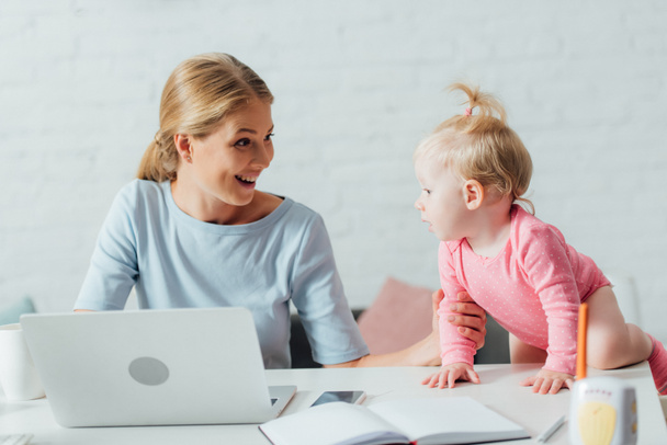 Szelektív fókusz a nő nézi a lányát, miközben dolgozik laptop közelében baby monitor az asztalon  - Fotó, kép