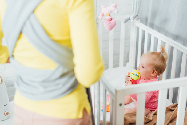 Selective focus of infant girl biting toy in crib near mother with sling at home  - Photo, Image
