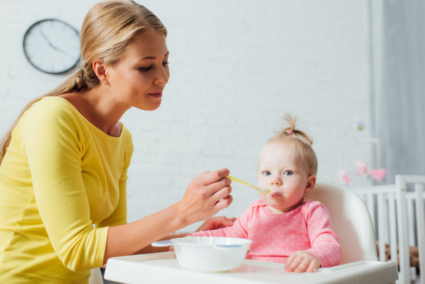 Woman holding spoon of baby food while feeding daughter on highchair at home  - Foto, afbeelding
